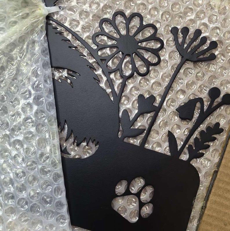 Op de afbeelding: Een matzwart metalen wanddecoratie met het silhouet van een hondenkop versierd met diverse bloemstengels, waaronder een grote margriet en een takje met bessen, met een uitgesneden pootafdruk aan de onderkant van het ontwerp.