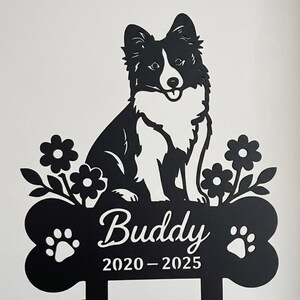 Op de afbeelding: Een zwart metalen bord met een silhouet van een hond die boven een botvormige basis zit. Op het bot staan de naam Buddy en de jaren 2020-2025. Twee pootafdrukken flankeren de tekst en bloemmotieven bevinden zich aan beide zijden van de hond.