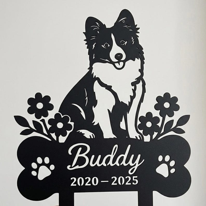 Op de afbeelding: Een zwart metalen bord met een silhouet van een hond die boven een botvormige basis zit. Op het bot staan de naam Buddy en de jaren 2020-2025. Twee pootafdrukken flankeren de tekst en bloemmotieven bevinden zich aan beide zijden van de hond.