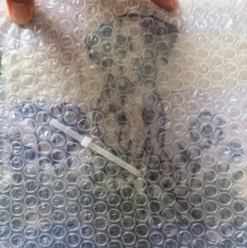 Op de afbeelding: Een doorzichtige plastic noppenfolie verpakking met een donkere, onduidelijke grafische illustratie van een hond. Een dunne, witte plastic tie-wrap is diagonaal over het midden van het oppervlak van de noppenfolie bevestigd.