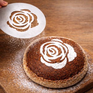 Puede incluir: Una hogaza de pan redonda con un diseño de rosa blanca espolvoreado en la parte superior. Se sostiene una plantilla con un diseño de rosa sobre el pan, con harina cayendo sobre la hogaza. El pan está sobre una tabla de cortar de madera.