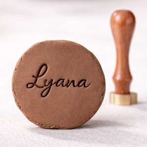 May include: A brown, circular stamp with the name "Lyana" elegantly engraved in cursive script. A wooden handle with a brass base is positioned nearby. The stamp is likely used for crafts or personalization.