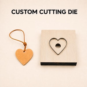 以下が含まれることがあります： 「CUSTOM CUTTING DIE」という文字が書かれたカスタムカッティングダイ。正方形の木製ブロックにはハート型の金属刃と円形の穴があり、その横には革製のハート型タグと、同じ革製の紐のループが添えられています。