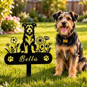 Op de afbeelding: Een zwarte metalen tuinpaal met een silhouet van een ruwharige hond zittend tussen bloemen, met de naam Bella in schrijfletter en twee pootafdrukken op een rechthoekige basis, naast een echte hond met een bruine halsband.