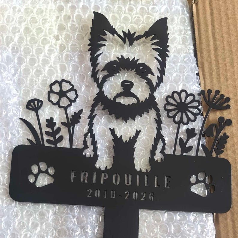 Op de afbeelding: Een zwarte metalen tuinpaal met een silhouet van een terri&euml;r hond omringd door diverse bloemen. De rechthoekige basis bevat de naam FRIPOUILLE, de jaren 2010-2026 en twee uitgesneden pootafdrukken aan de linker- en rechterkant.