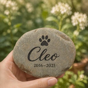 Op de afbeelding: Een gladde, ovaalvormige grijze gedenksteen met een gegraveerde zwarte pootafdruk, de naam Cleo in een cursief lettertype en de jaartallen 2016-2025. De steen heeft een natuurlijk, getextureerd oppervlak met zichtbare variaties in kleur en tint.
