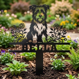 Op de afbeelding: Een zwarte metalen tuinpaal met een silhouet van een ruwharige hond die tussen twee bloemstukken zit. De naam BELLA is uitgesneden in dikke hoofdletters op een horizontale balk onder de hond, ondersteund door een verticale paal.