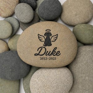 May include: A smooth, tan-coloured river stone featuring a black laser-engraved graphic of a dog silhouette with angel wings and a halo above its head. Below the graphic, the name Duke is engraved in script, followed by the dates 2012-2025.