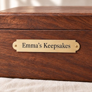 Può includere: Una scatola rettangolare in legno marrone scuro con una placca orizzontale in ottone spazzolato fissata con due piccole viti. La placca presenta l'incisione del testo nero "Emma's Keepsakes" in un carattere serif, centrata sul pannello.