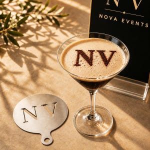 Op de afbeelding: Een rond, plat, zilverkleurig metalen sjabloon met de uitgesneden letters NV, geplaatst naast een martiniglas gevuld met een donkere vloeistof met een schuimlaag. Het schuim toont de letters NV in donker poeder, passend bij het sjabloon.
