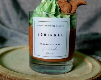 Könnte beinhalten: Eine handgemachte Kerze aus Bio-Sojawachs in einem klaren Glas. Die Kerze hat eine braune Basis, gekrönt von grünem Wachs und einer braunen Eichhörnchenfigur. Das Etikett lautet "Squirrel". Die Kerze ist 226g.