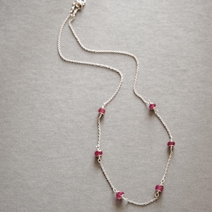 May include: A delicate silver necklace featuring small, vibrant red gemstones evenly spaced along the chain. The necklace has a simple, elegant design, perfect for everyday wear. The gemstones are a deep, rich red color, adding a pop of color to the silver chain.