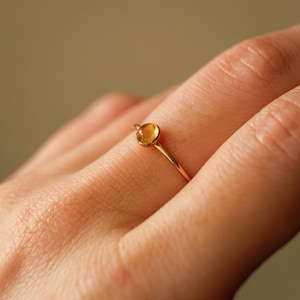May include: A delicate gold ring featuring a small, round, amber-colored gemstone. The ring has a thin, polished gold band. The gemstone is set in a simple bezel setting, creating a minimalist and elegant design. The ring is shown on a person's finger.