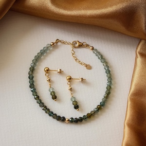 Puede incluir: Un conjunto de pulsera y pendientes dorados. La pulsera y los pendientes presentan pequeñas cuentas facetadas de color verde y verde azulado. Los pendientes tienen un diseño colgante con un poste dorado y pequeñas cuentas.