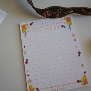 以下が含まれることがあります： 金色の水平線と花のイラストが入った白いメモ帳。「Across the Stars」という文字が上部に金色の筆記体で書かれています。メモ帳の下部には「Sounds an awful lot like a dictatorship to me. Well, if it works.」という文字が書かれています。