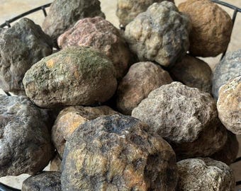 Pode incluir: Uma coleção de grandes geodos redondos de cor cinza, marrom e verde em uma cesta de arame preto. Os geodos têm uma superfície áspera e texturizada.