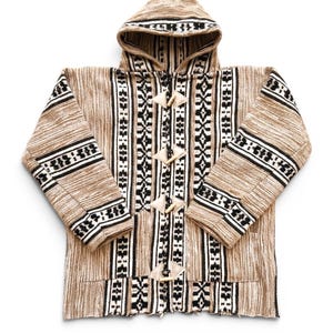 May include: A hooded jacket featuring a tan, black, and white woven pattern with vertical stripes and geometric motifs. The front closure includes four light-coloured wooden toggles with fabric loops. The text "Marrakech craft work" is below.