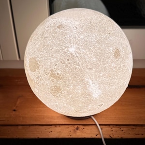 Könnte beinhalten: Eine leuchtende, kugelförmige Lampe, die dem Mond ähnelt, mit einer strukturierten Oberfläche und komplizierten Details. Die Lampe ist weiß und steht auf einem dunklen Sockel, mit einem weißen Netzkabel. Die Lampe steht auf einer Holzoberfläche.