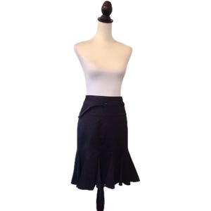 May include: A black skirt with a flared hemline, displayed on a mannequin. The skirt features a belt with a buckle and a unique asymmetrical design element on the waist. The hem has a ruffled, layered appearance.