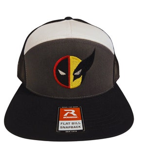 Puede incluir: Una gorra de camionero negra, gris y blanca con una parte trasera de malla negra. La gorra tiene una visera plana y un cierre a presión. La parte delantera de la gorra presenta un logotipo bordado de la cabeza de Wolverine, con un esquema de color amarillo y rojo. La gorra también tiene una pequeña etiqueta negra y naranja que dice "Richardson" y "Flat Bill Snapback".