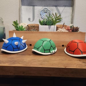 Puede incluir: Tres réplicas de caparazones de tortuga de colores azul, verde y rojo, cada una con un borde blanco. El caparazón azul tiene púas blancas. Se exhiben sobre una superficie de madera, lo que sugiere un tema de juego o novedad.