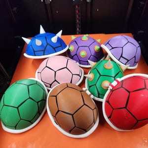 May include: A collection of colorful, 3D-printed turtle shells in various colors, including blue, purple, pink, green, brown, and red. Each shell features a geometric pattern and white trim. Some shells have white spikes.