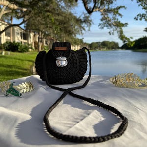 May include: A black crocheted handbag with a silver clasp and a long strap. The bag is displayed with two gold-colored leaf crowns and a gold and green leaf bracelet. The items are on a white surface with a lake and trees in the background.