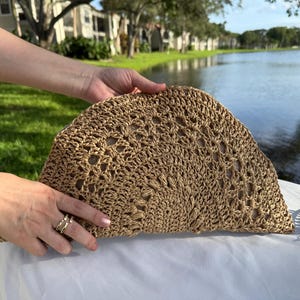 May include: A woven, half-moon shaped clutch in a light brown color. The clutch has an open weave design and is held by a person. A matching bracelet is also visible. The background includes a lake and green grass.