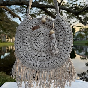 May include: A beige, round crochet bag with a long strap. The bag features a detailed circular pattern, wooden beads, and a tassel. The bottom edge has long fringe. The bag is hanging in front of a blurred outdoor background.