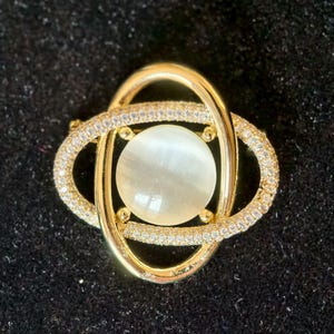 May include: A gold-toned brooch with a central, round, opalescent stone. The brooch features an oval frame encrusted with small, clear crystals, and is intertwined with a gold-colored design. The background is a dark, textured surface.