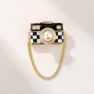以下が含まれることがあります： 黒と金のカメラ型のブローチ。レンズはパールとラインストーン。カメラ本体は白黒の市松模様で、金のチェーンが付いています。ブローチには長方形のラインストーンと小さな丸いラインストーンが付いています。