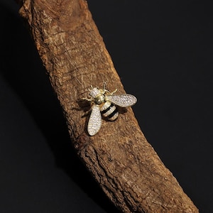 Puede incluir: Un broche de abeja dorada con esmalte negro y diamantes de imitación transparentes. La abeja está posada sobre un trozo de corteza marrón.