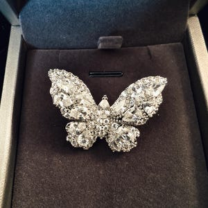 May include: A silver butterfly brooch, set with clear gemstones, is displayed in a dark grey jewellery box. The brooch's wings are detailed with multiple stones, creating a sparkling effect. The box has a plush, velvety interior.