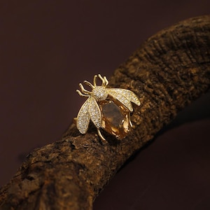 Pode incluir: Um anel de ouro com um design de abelha. A abelha é incrustada com pedras transparentes e tem uma pedra marrom no centro.