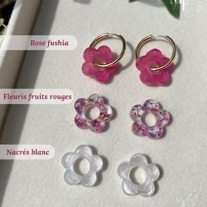 Puede incluir: Una selección de pendientes con forma de flor en varios colores. El par superior presenta flores fucsia en aros dorados. Debajo hay dos pares de pendientes de flores, uno con detalles de frutos rojos y florales, y el otro en blanco nacarado.