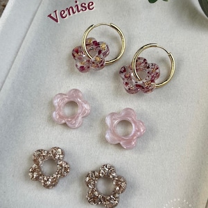 May include: Gold hoop earrings with flower-shaped pendants. The pendants are made of clear resin with red and gold accents. Additional flower-shaped resin pieces in pink and gold are also displayed. The word "Venise" is written in the upper left corner.