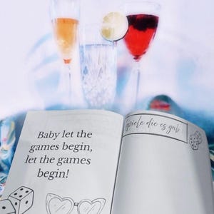 K&ouml;nnte beinhalten: Offenes Buch mit dem Text "Baby let the games begin, let the games begin!" und "spiele die es gab" auf den Seiten. Es sind zwei W&uuml;rfel und eine Sonnenbrille auf den Seiten gezeichnet.