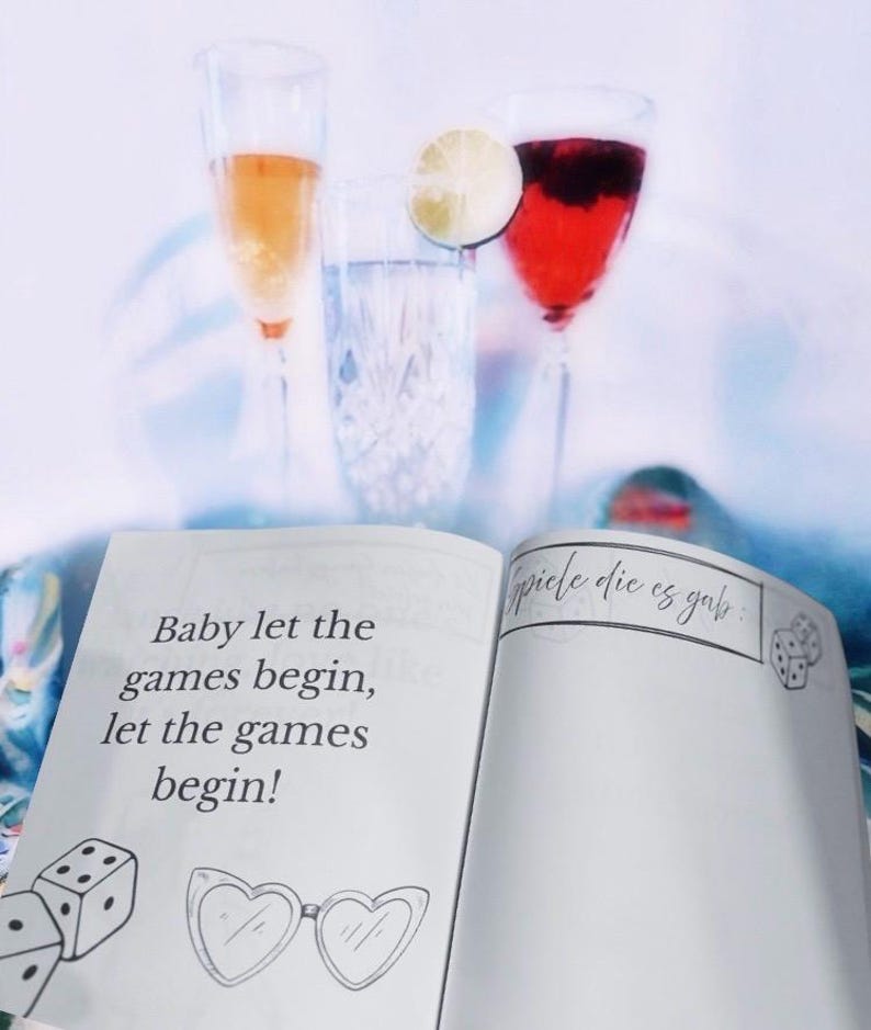 K&ouml;nnte beinhalten: Offenes Buch mit dem Text "Baby let the games begin, let the games begin!" und "spiele die es gab" auf den Seiten. Es sind zwei W&uuml;rfel und eine Sonnenbrille auf den Seiten gezeichnet.