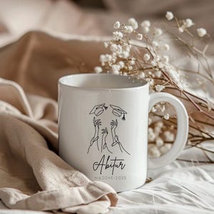 May include: White ceramic mug with a graduation design. The mug displays a black line drawing of hands throwing graduation caps, with the word "Abitur" and the name "NADINE 2025" in a script font. The mug has a large handle.