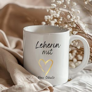 May include: White ceramic mug with black text that reads "Lehrerin mit" and a gold heart. Below the heart is the text "Frau Schulz".