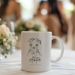 May include: White ceramic mug with a black line drawing of graduation caps and hands. The word "Abitur" is written in script, with "NADINE 2025" below. The mug has a curved handle.