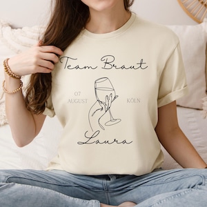 Op de afbeelding: Een crèmekleurig T-shirt met de tekst "Team Braut" in zwarte cursieve letters. Onder de tekst staat een lijntekening van een hand die een wijnglas vasthoudt. De datum "07 augustus" en het woord "Köln" staan onder het glas gedrukt. De naam "Laura" staat in zwarte cursieve letters onder het glas.