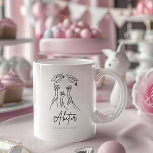 May include: White ceramic mug with a graduation theme. The mug features a black line drawing of hands tossing graduation caps, with the word "Abitur" and the name "NADINE-2025" printed below. The mug has a curved handle.