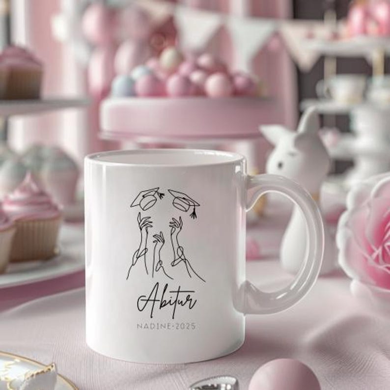 May include: White ceramic mug with a graduation theme. The mug features a black line drawing of hands tossing graduation caps, with the word "Abitur" and the name "NADINE-2025" printed below. The mug has a curved handle.