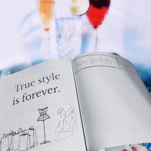 K&ouml;nnte beinhalten: Ein offenes Notizbuch mit dem Text "True style is forever." auf der linken Seite. Die rechte Seite hat einen handschriftlichen Titel "Choose Your Outfits" und ist leer. Das Notizbuch liegt auf einem Tisch mit zwei Gl&auml;sern Getr&auml;nken und einer Limettenscheibe im Hintergrund.