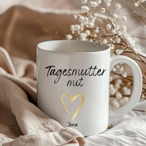 May include: White ceramic mug with black text that reads "Tagesmutter mit" and a gold heart with the name "Jana" underneath.