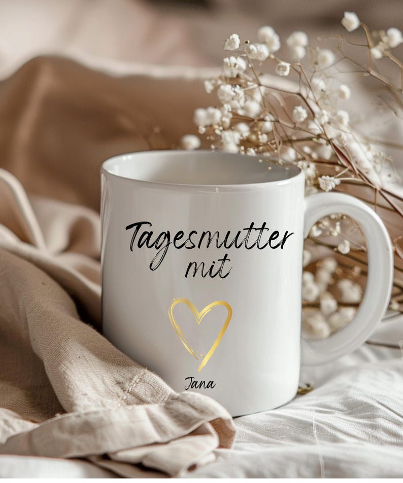 May include: White ceramic mug with black text that reads "Tagesmutter mit" and a gold heart with the name "Jana" underneath.