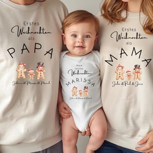 May include: A family wearing matching beige sweatshirts and a white onesie. The sweatshirts say "Erstes Weihnachten als PAPA" and "Erstes Weihnachten als MAMA" with gingerbread family graphics. The onesie says "Mein erstes Weihnachten MARISSA".