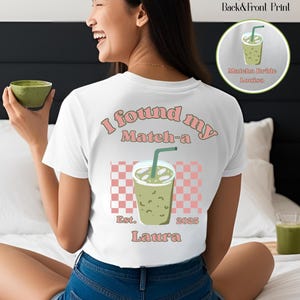 May include: White t-shirt with a graphic of a matcha drink and the text "I found my Match-a Est. 2025 Laura". The design includes a pink and white checkered pattern. A small image of a matcha drink is in the upper right corner.