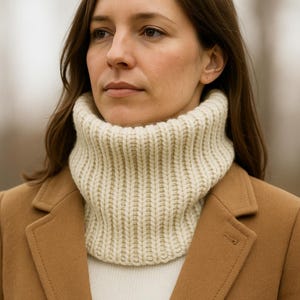May include: A cream-colored, ribbed knit cowl neck warmer. The neck warmer is worn over a white sweater and under a tan coat. The cowl is made of a thick, soft yarn and has a high, folded collar.